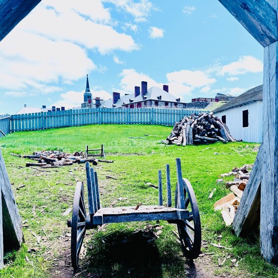 A historical site with a wooden fence, a pile of logs, and a cart in the foreground. 3
