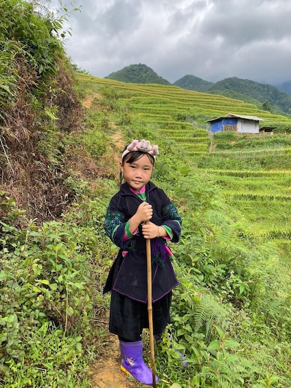 A child in traditional attire standing in a lush green rice paddy field, holding a wooden stick. 3