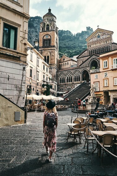 A cobblestone street with a person walking, historic buildings, and a bell tower. 1