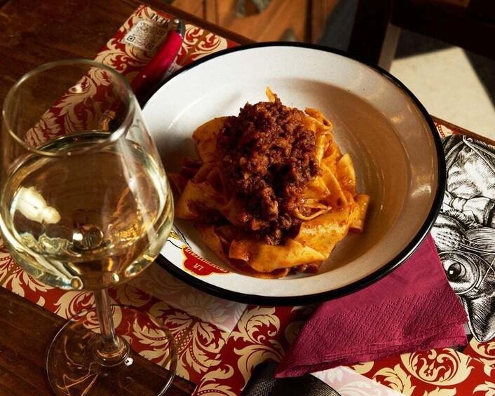 A plate of pasta with a meat topping, a glass of white wine, and a patterned tablecloth. 3