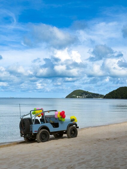 A blue off-road vehicle with colorful balloons parked on a sandy beach, with a calm sea and a hilly island in the background. 2