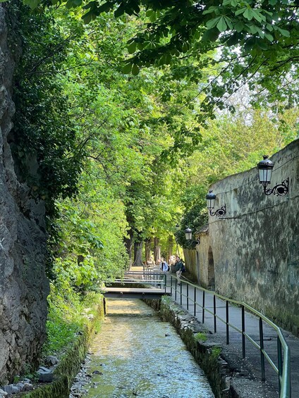 A narrow waterway flanked by stone walls and lush greenery, with a pedestrian path and street lamps. 3