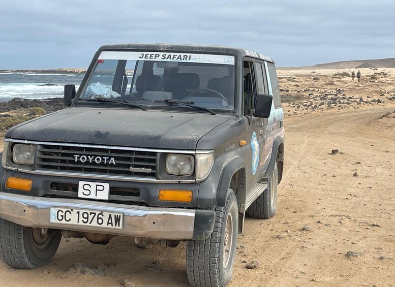 A Toyota SUV with 'JEEP SAFARI' on the windshield, parked on a sandy beach with rocky outcrops in the background. 4