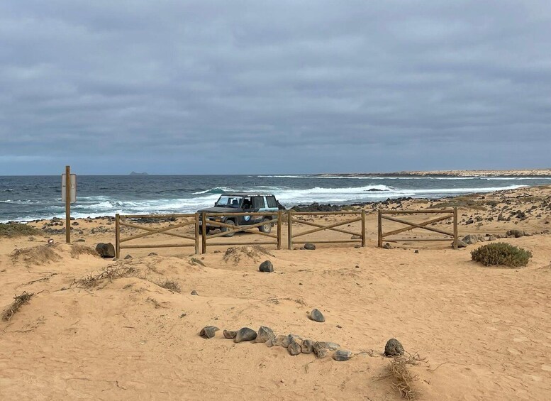 A beach with a vehicle parked near a wooden gate, ocean waves, and a cloudy sky. 1