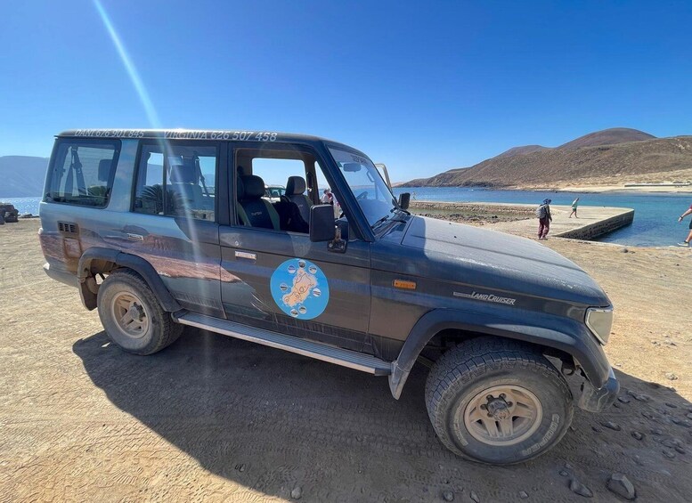 A dark-colored SUV with a 'Land Cruiser' badge parked on a sandy beach with a clear blue sky and mountains in the background. 3