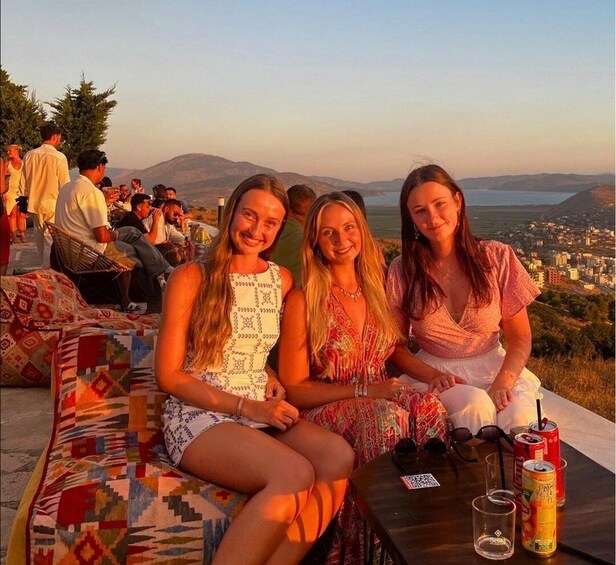 Three women sitting on a patterned couch with a scenic view of a city and mountains in the background. 1