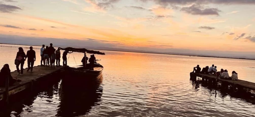 A sunset over a calm body of water with people on a dock and a boat. 1