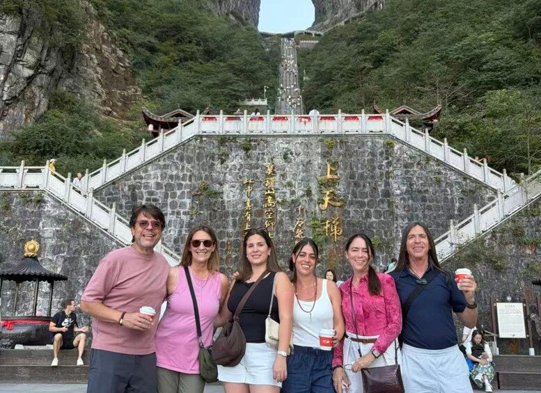 A group of people standing in front of a stone monument with inscriptions, a staircase, and a mountainous backdrop. 2