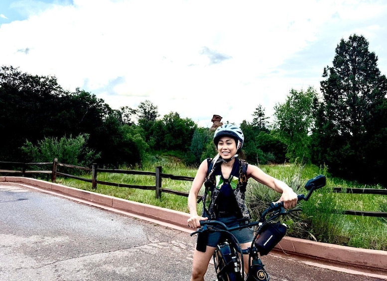 A person on a bicycle with a helmet, wearing a backpack, and a water bottle on the bike, standing on a paved path with grass and trees in the background. 4