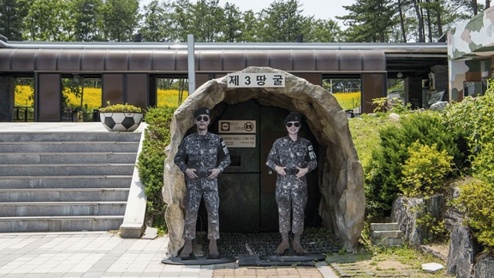 Two soldiers in camouflage uniforms standing in front of a stone structure with a sign above it. 4