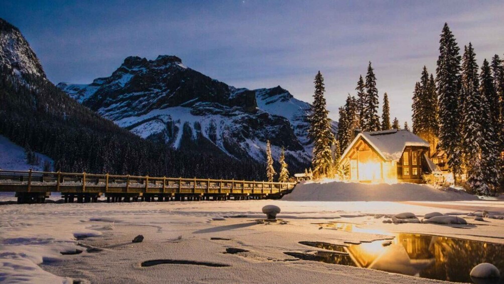 A snow-covered landscape with a wooden cabin, a wooden bridge, and a mountain range in the background. 2