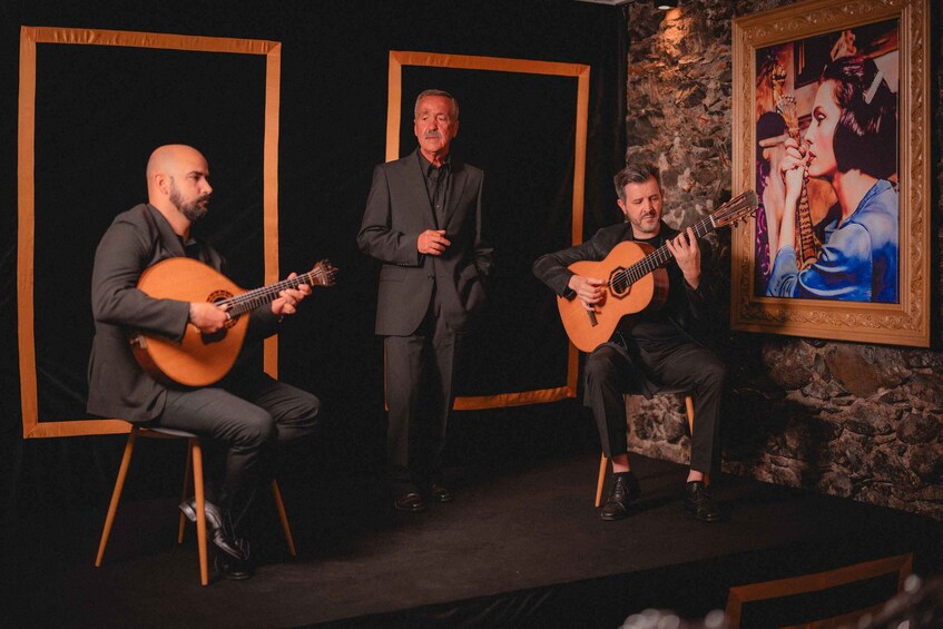 Three men performing on stage with guitars, a framed painting, and a stone wall in the background. 3