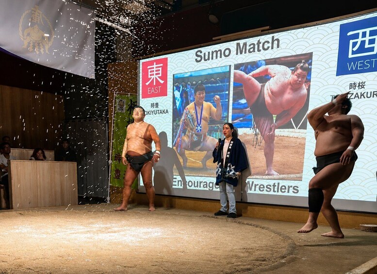 A sumo wrestling match with a large audience, a stage with a backdrop displaying 'Sumo Match' and 'East', and a person speaking on stage. 3