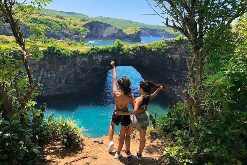 Two people standing on a dirt path overlooking a blue water body with a rocky cliff and greenery. 2
