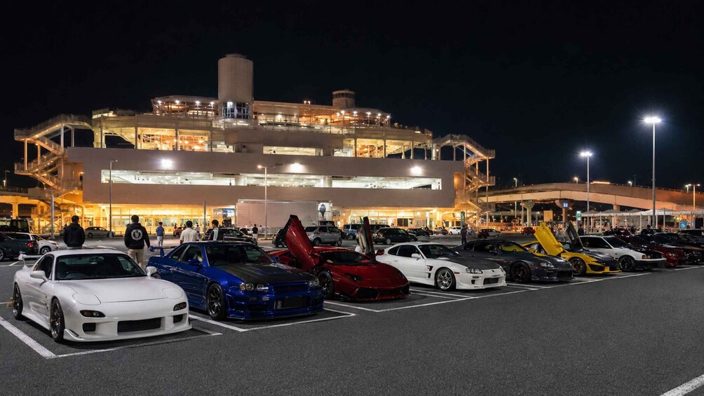 A parking lot with several sports cars and a large building with multiple levels and lights. 1