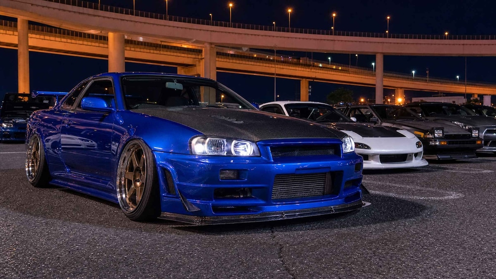A blue sports car with a black hood parked in a lot with other cars and a bridge in the background. 3