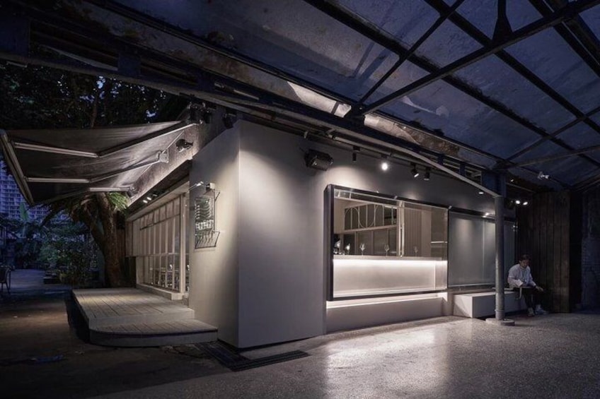 A modern, illuminated outdoor kiosk with a glass enclosure, a wooden bench, and a metal roof structure. 2