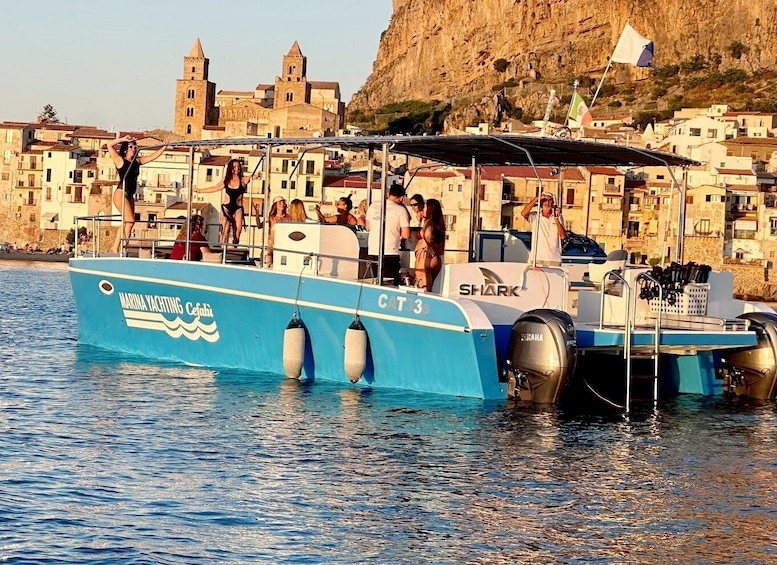 A blue boat with 'MARINA YACHTING CAFFO' and 'CT3' on its side, docked near a coastal town with buildings and cliffs in the background. 2