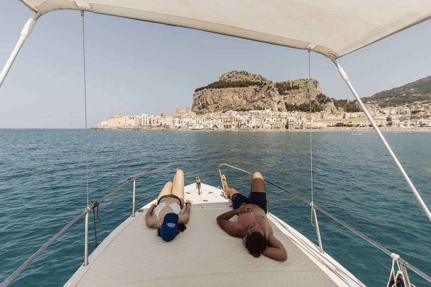 Two people relaxing on a boat with a coastal town and a prominent hill in the background. 2