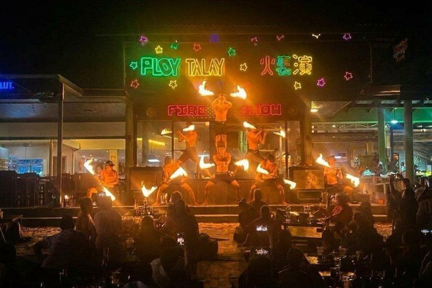 A nighttime outdoor performance with performers on a stage, illuminated by bright lights and fire effects. 2