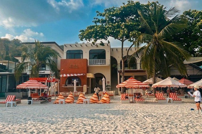 A beach with lounge chairs and umbrellas, palm trees, and a building with a sign. 5