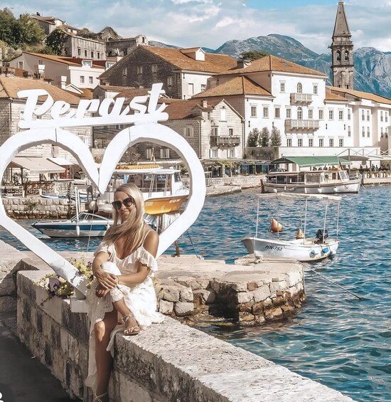 A woman in a white dress sitting by a waterfront with a large heart-shaped sign reading 'Perist.' 3