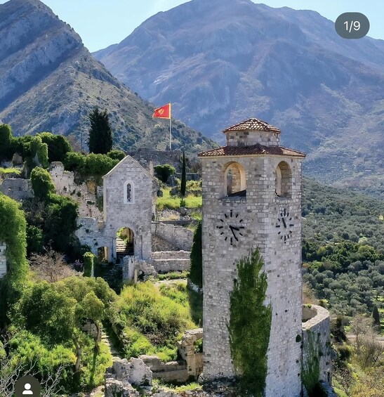 A historic stone tower with a clock, surrounded by lush greenery and a flag atop a nearby structure. 2