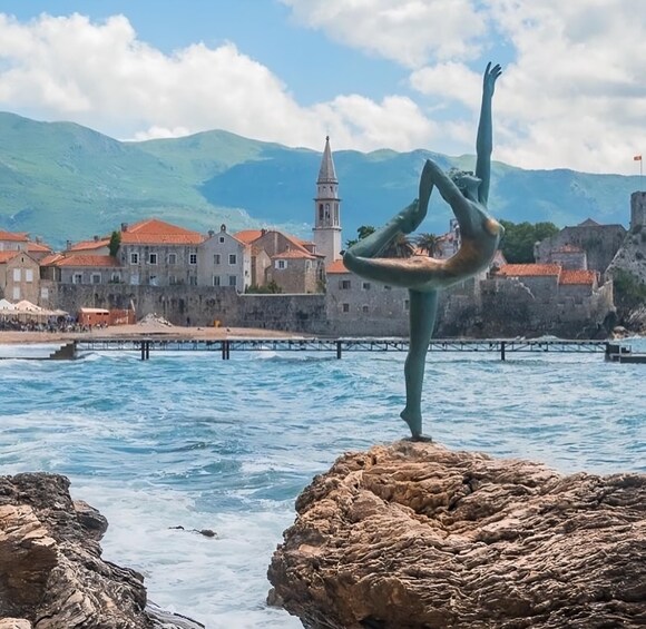 A statue of a person performing a yoga pose on a rock by a body of water with buildings and mountains in the background. 1