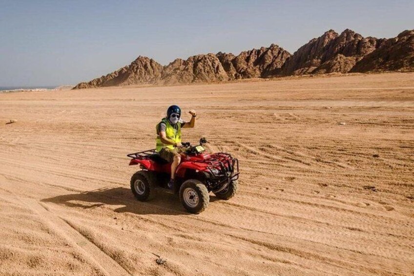 A person riding a red ATV in a desert with sand dunes and rocky mountains in the background. 5