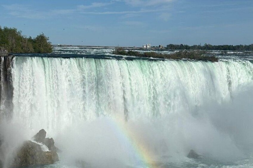 A large waterfall with a rainbow, surrounded by trees and buildings in the background. 1