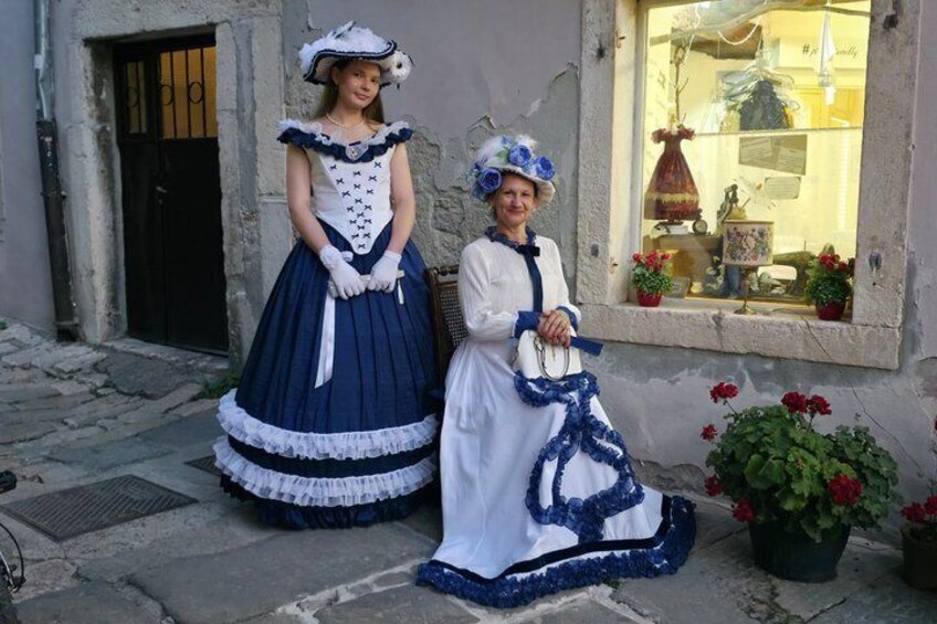 Two individuals in historical costumes standing in front of a building with a window displaying flowers. 5