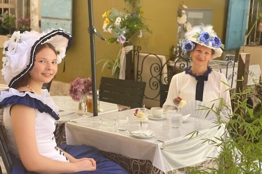 Two people seated at an outdoor table with a white tablecloth, surrounded by greenery and a yellow wall. 4