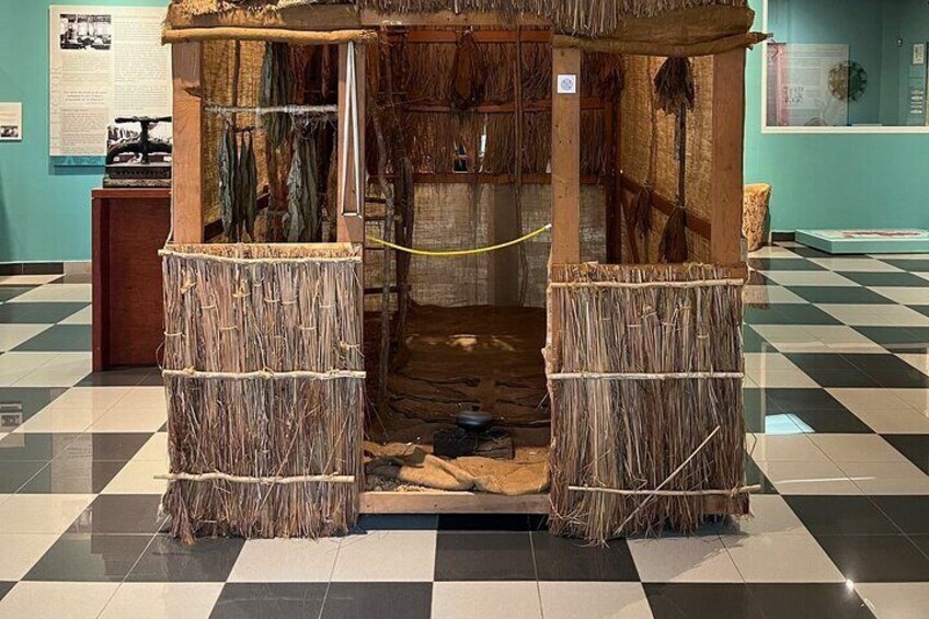 A traditional hut with thatched walls and a wooden door, displayed in a museum setting. 1