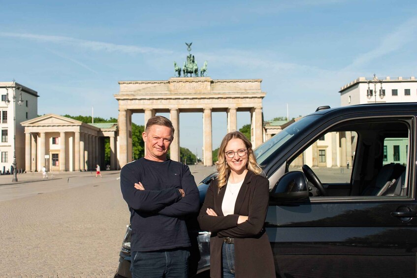 Two people standing in front of a black car with the Brandenburg Gate in the background. 1