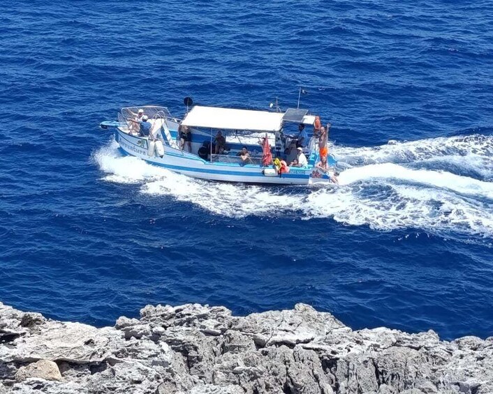A boat with passengers on a clear blue sea, moving away from rocky shore. 5