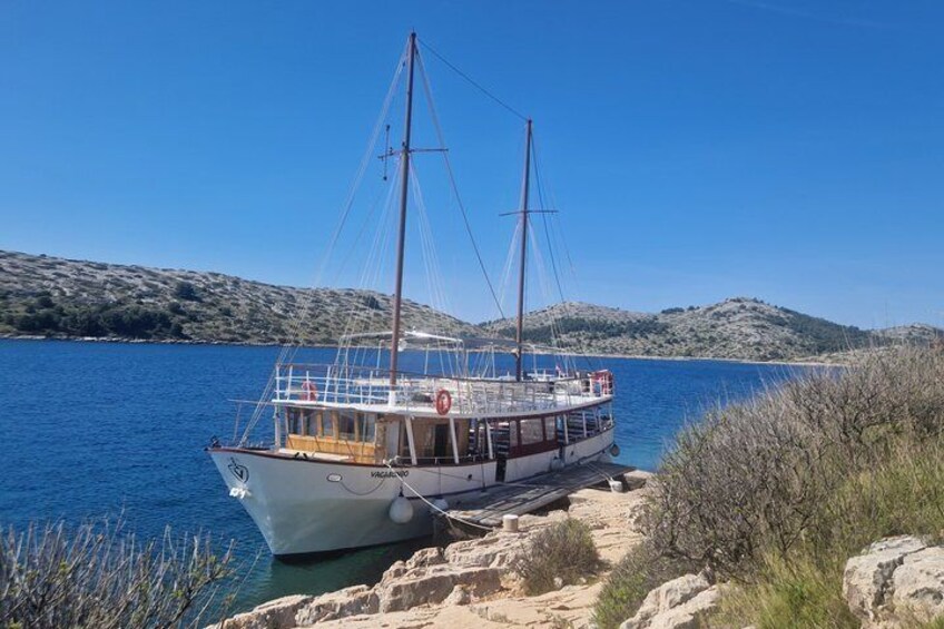 A boat docked at a rocky shore with clear blue water and hills in the background. 1