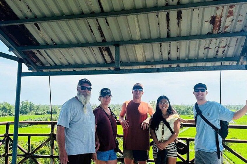 A group of people standing under a metal roofed structure with a green field in the background. 5