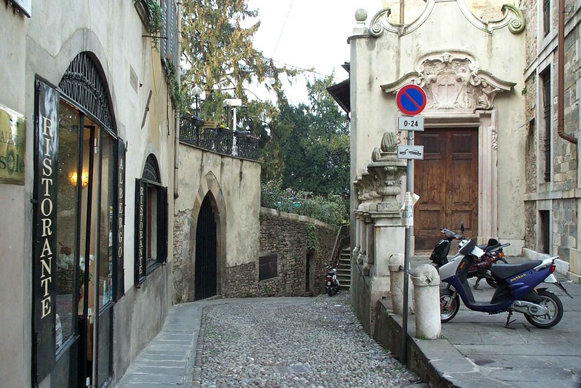 A narrow cobblestone street with a restaurant named 'RISTORANTE' on the left, a no parking sign, and a scooter parked on the right. 5
