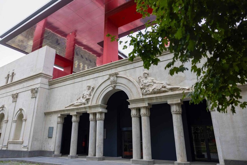 Ein klassizistisches Gebäude mit Säulen und Statuen, ein rotes Dach und ein Baum im Vordergrund. 3