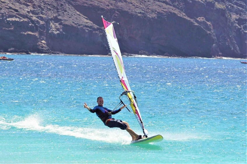 A person windsurfing on a clear blue sea with a mountainous coastline in the background. 1