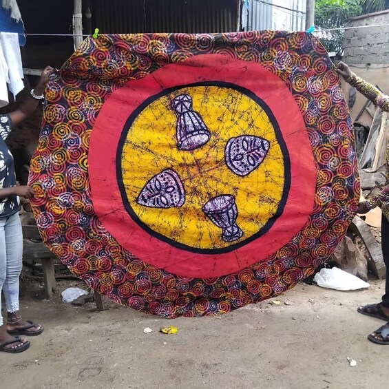 A colorful tapestry with a central yellow circle and four purple objects, displayed outdoors on a concrete surface. 3
