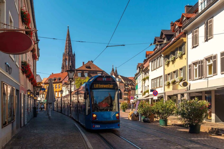 A tram labeled 'Littenweiler' on a cobblestone street with historic buildings and a church spire in the background. 3