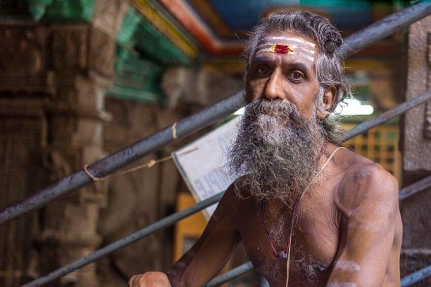 A person with a long beard and a red bindi on their forehead, standing in front of a stone structure with metal bars. 4