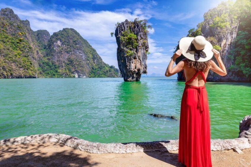 A woman in a red dress and white hat standing by a turquoise sea with rocky cliffs. 1