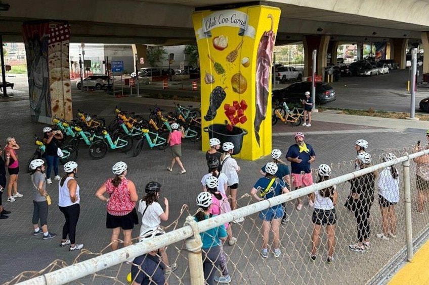 A group of people with bicycles near a yellow pillar with a mural. 1