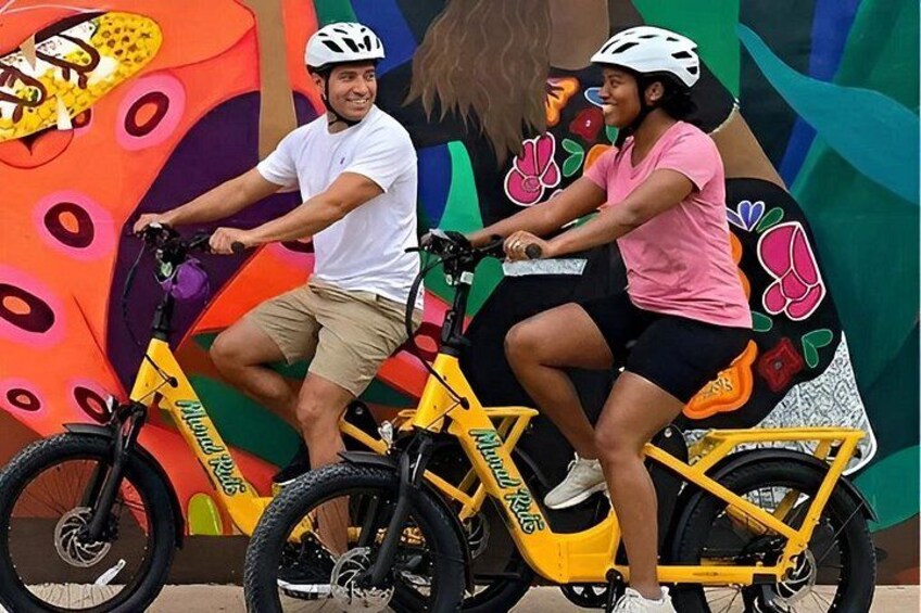 Two people riding yellow bicycles with a colorful mural in the background. 4