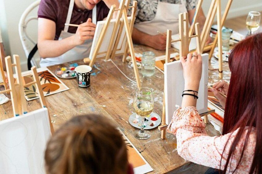 A group of people engaged in painting on easels at a wooden table. 4