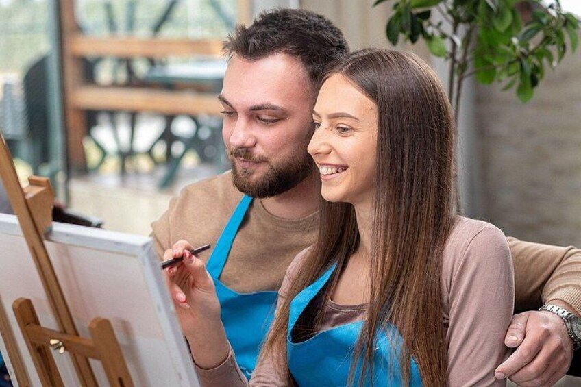 A person painting on a canvas with an easel, another person observing, and a plant in the background. 5
