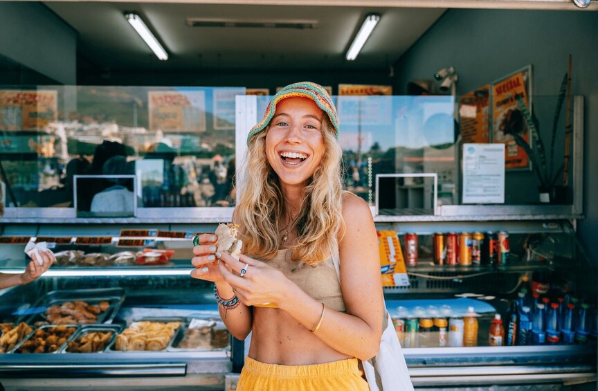 A person in a yellow top and striped hat holding a sandwich at a food stall with various food items and condiments visible. 3