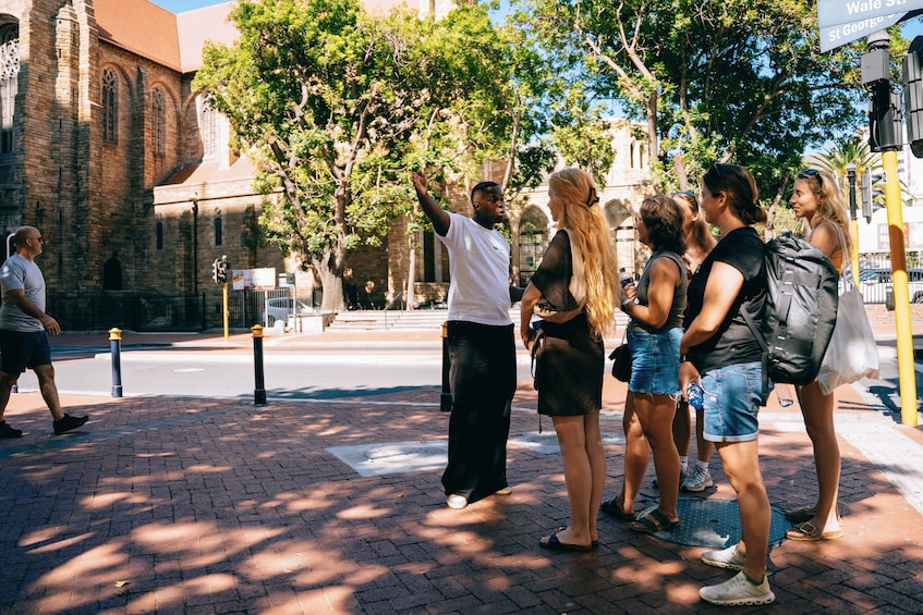 A group of people standing on a brick-paved sidewalk near a historic building with arched windows and a clock tower. 2